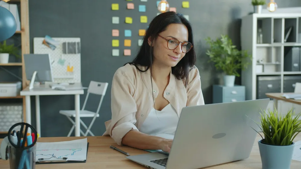 A professional smiling at her laptop in a modern office, representing a career changer landing a new role after retraining
