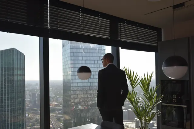 A founder in a dark suit stands at a floor-to-ceiling window overlooking a city skyline, thinking through strategy before turning to AI tools